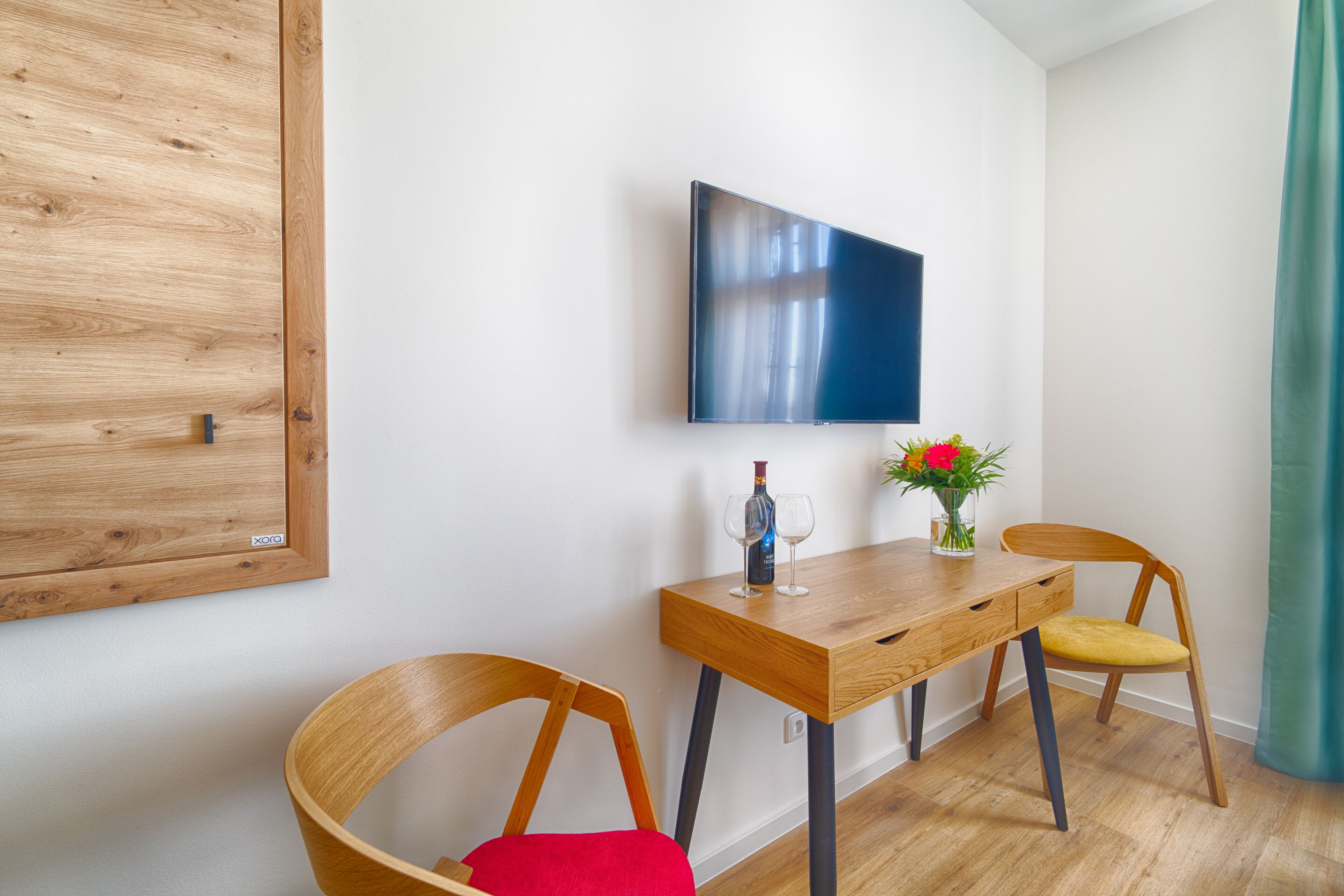 A modern room with a wooden desk, two wooden chairs, a flat-screen TV mounted on the wall, and a small table with a vase of flowers.