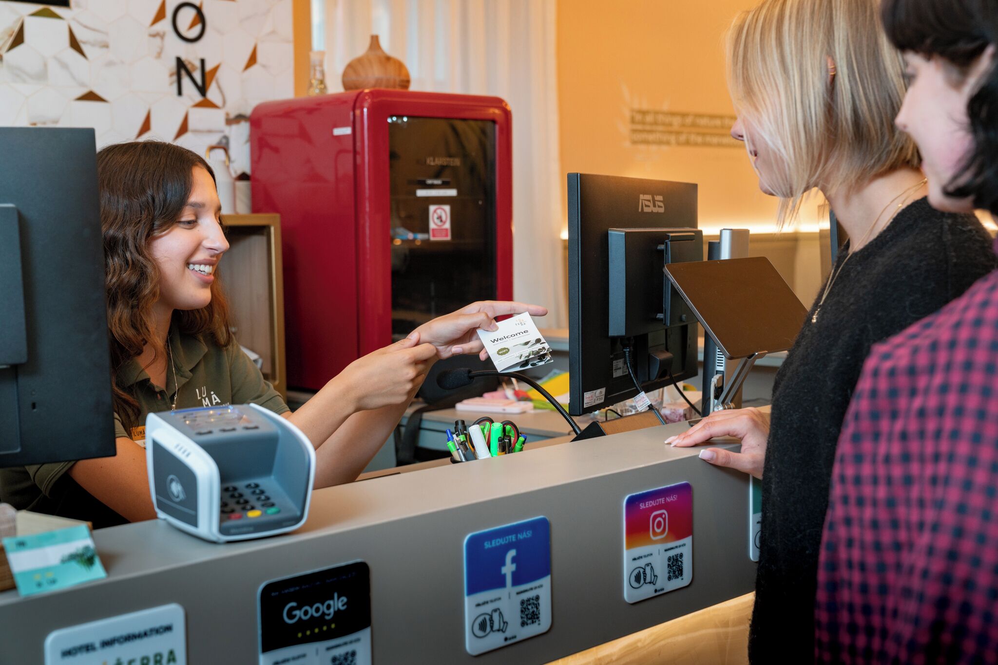 A reception desk with a digital payment terminal, a red refrigerator, and monitors.