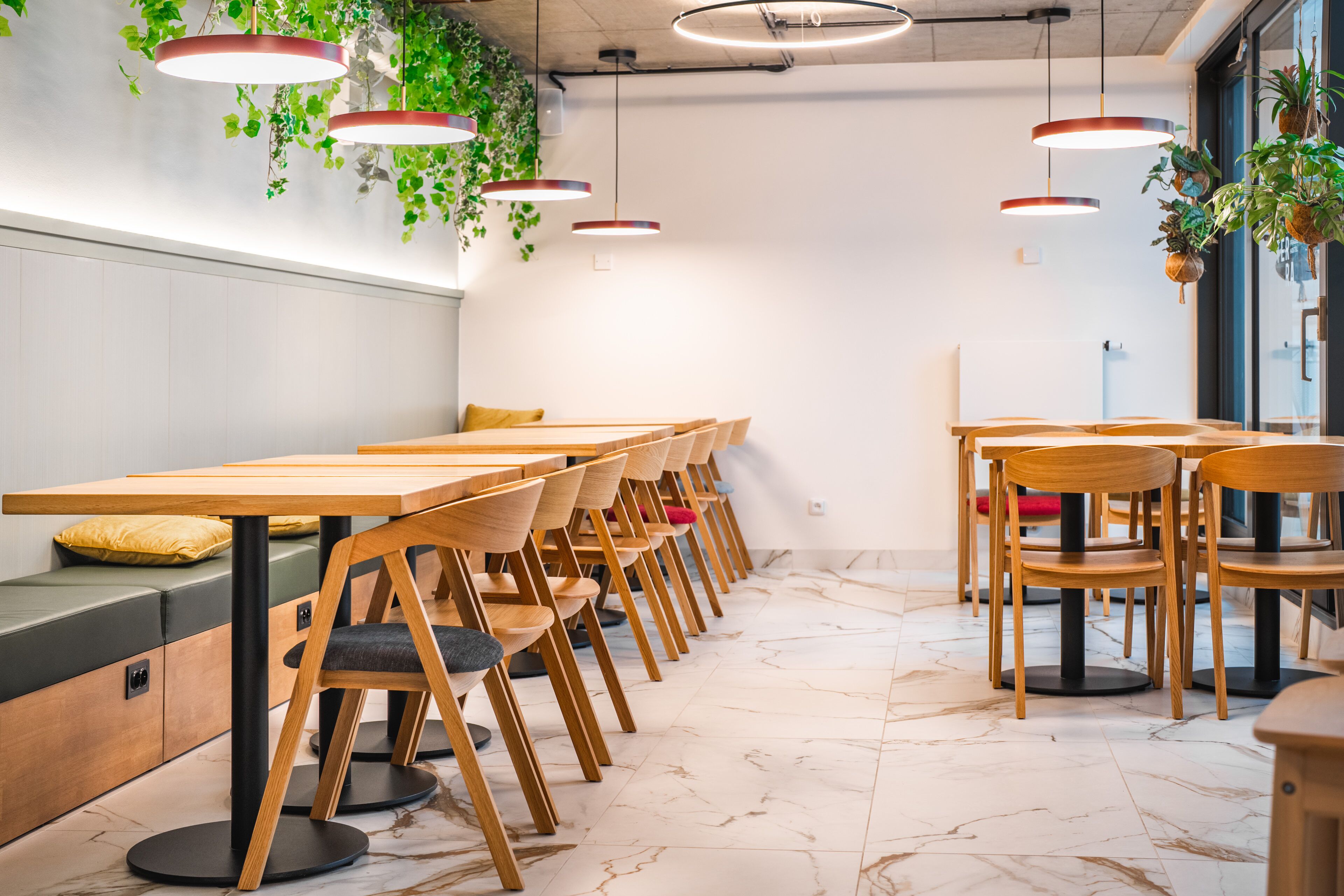 Un comedor moderno con mesas y sillas de madera, piso de mármol y plantas colgantes.