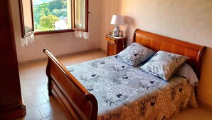 A bedroom with a wooden bed, a nightstand with a lamp, a window with curtains, and a view of a building and greenery outside. - Charming house near AJACCIO (Appietto)