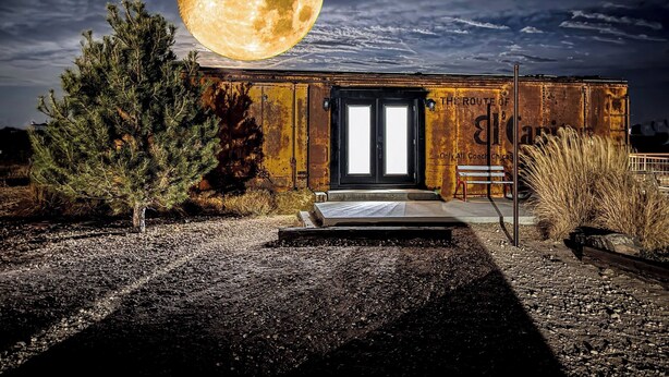 Un bâtiment rustique, avec une grande lune dans le ciel, une porte en métal et un banc à l’extérieur.