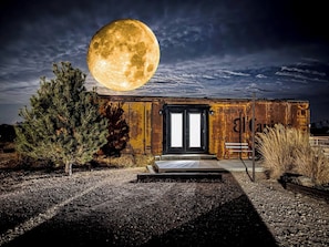 Un edificio rústico con una luna grande en el cielo, una puerta de metal y un banco afuera.