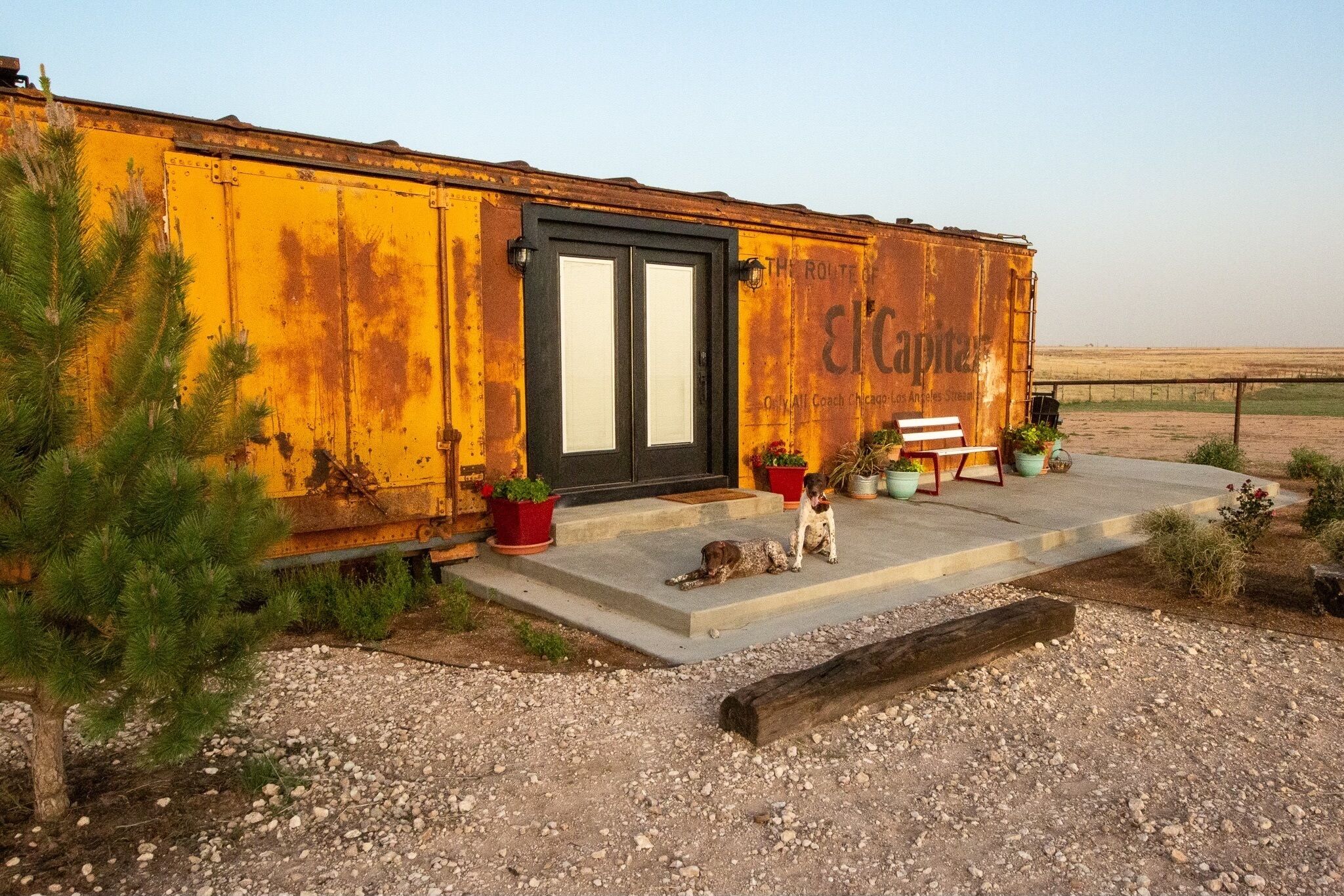 Un edificio rústico amarillo con una puerta negra, plantas en macetas y un banco.
