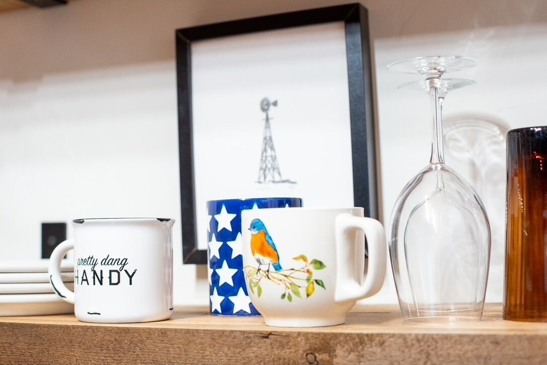 A shelf with various items including a mug with text, a mug with a bird design, a clear glass, and a framed picture.