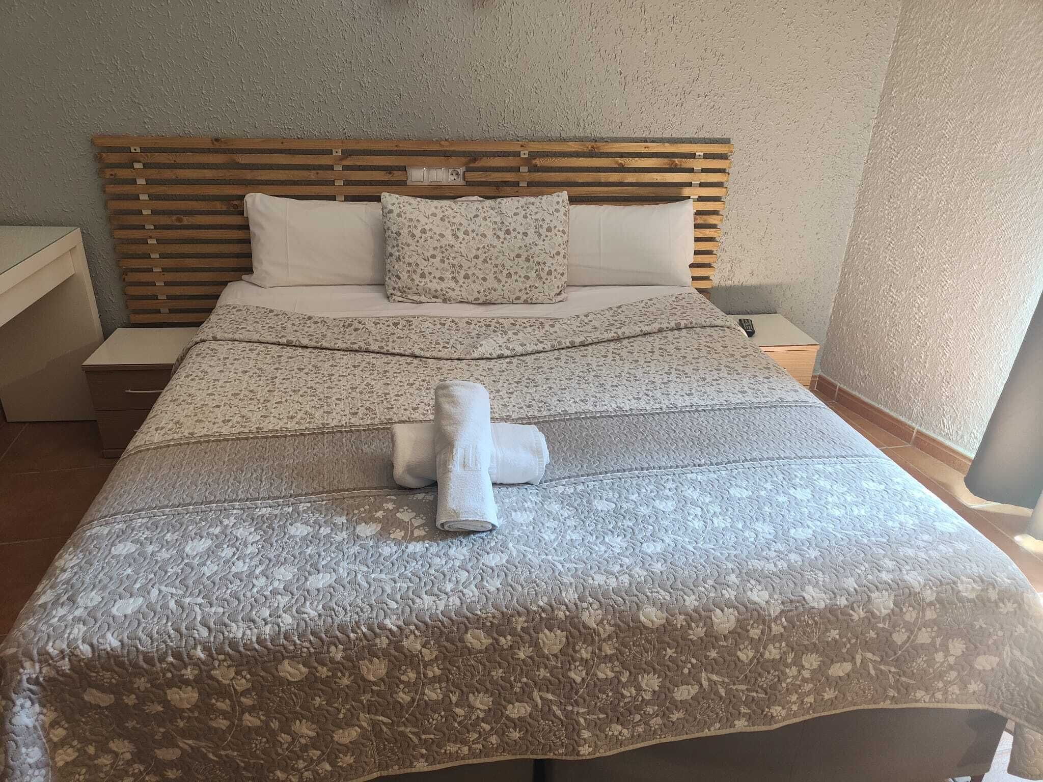 Una cama bien hecha con un edredón estampado con motivos florales, un cabecero de madera y un control remoto sobre la mesita de noche.