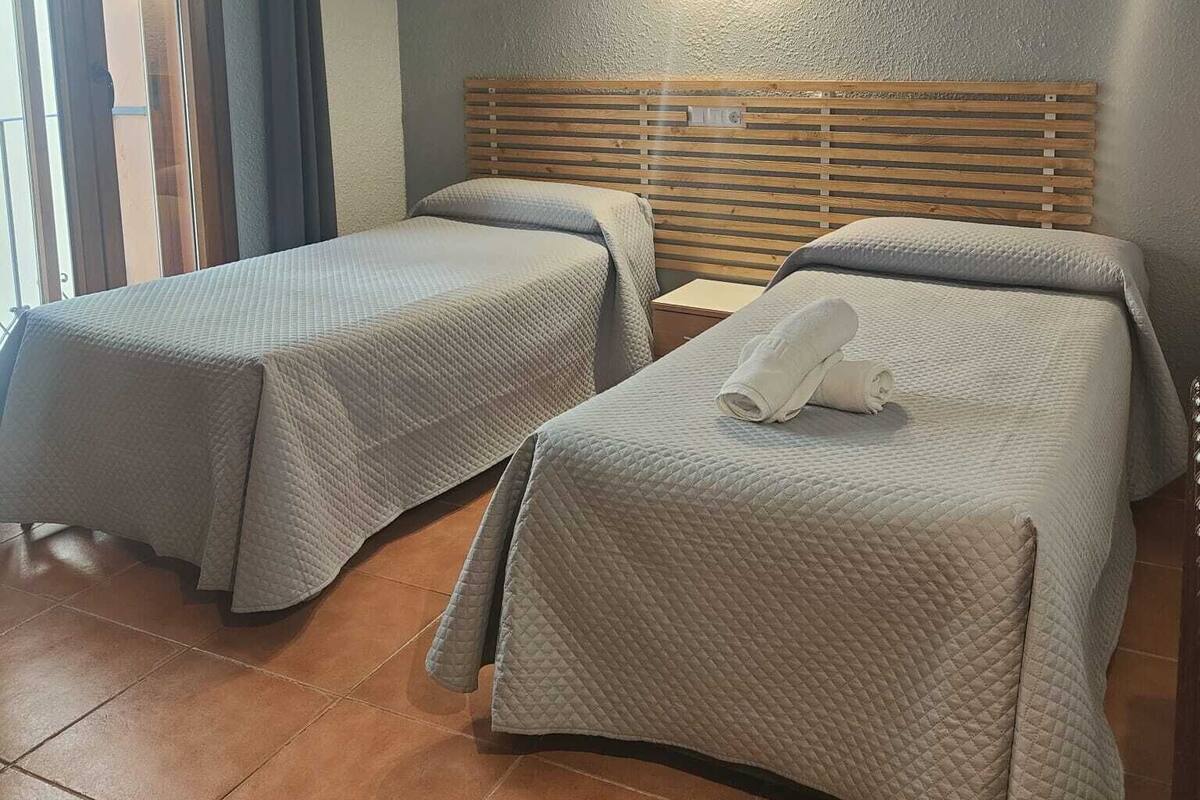 Habitación de hotel con dos camas, cabecero de madera, mesita de noche y ventana con cortinas.