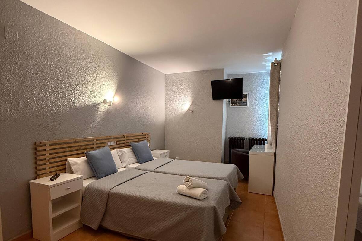 Habitación de hotel con dos camas, un televisor fijado a la pared y una mesita de noche pequeña con un control remoto.