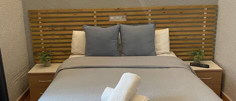 A neatly made bed with a quilted bedspread, two pillows, and a folded towel on the bed. There is a nightstand with a remote control and a small plant beside the bed.
