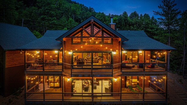 A two-story log cabin with a balcony on each floor, string lights, and a view of the surrounding forest.
