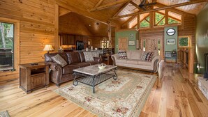 A spacious wooden cabin interior with a high ceiling, a dining area, and a living room furnished with leather sofas and a coffee table.