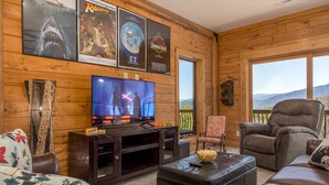A cozy living room with wooden walls, a large flat-screen TV, leather sofas, and a scenic mountain view through the window.