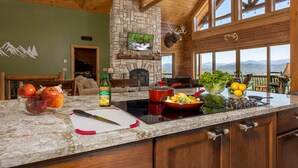 A kitchen with a stone fireplace, a large window with a mountain view, and a bowl of fruit on the counter.