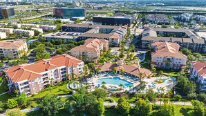 An aerial view of a resort with a central swimming pool, surrounded by multi-story buildings with red-tiled roofs, and a body of water in the foreground.