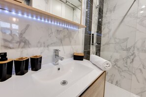 A modern bathroom with a white sink, black and wooden trash bins, a mirror with LED lights, and marble walls. - Les Mulots - Apartment T3 of 56 sqm (Annecy)
