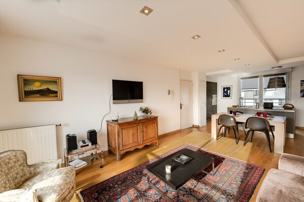 Una sala de estar con sofá, una mesa de centro y televisor. Hay un comedor con sillas y una mesa. El cuarto tiene piso de madera y paredes blancas.