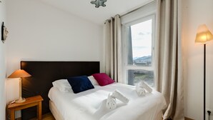 A neatly made bed with white linens, a wooden nightstand with a lamp, and a view of the outdoors through a window with curtains. - Le Kaliste*** - apartment for 4 people + garage (Annecy)