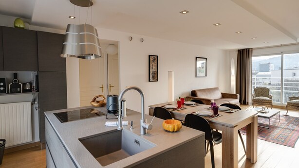 A modern kitchen with a central island, a dining area with a table and chairs, and a view of the city through large windows. - Le Kaliste*** - apartment for 4 people + garage (Annecy)