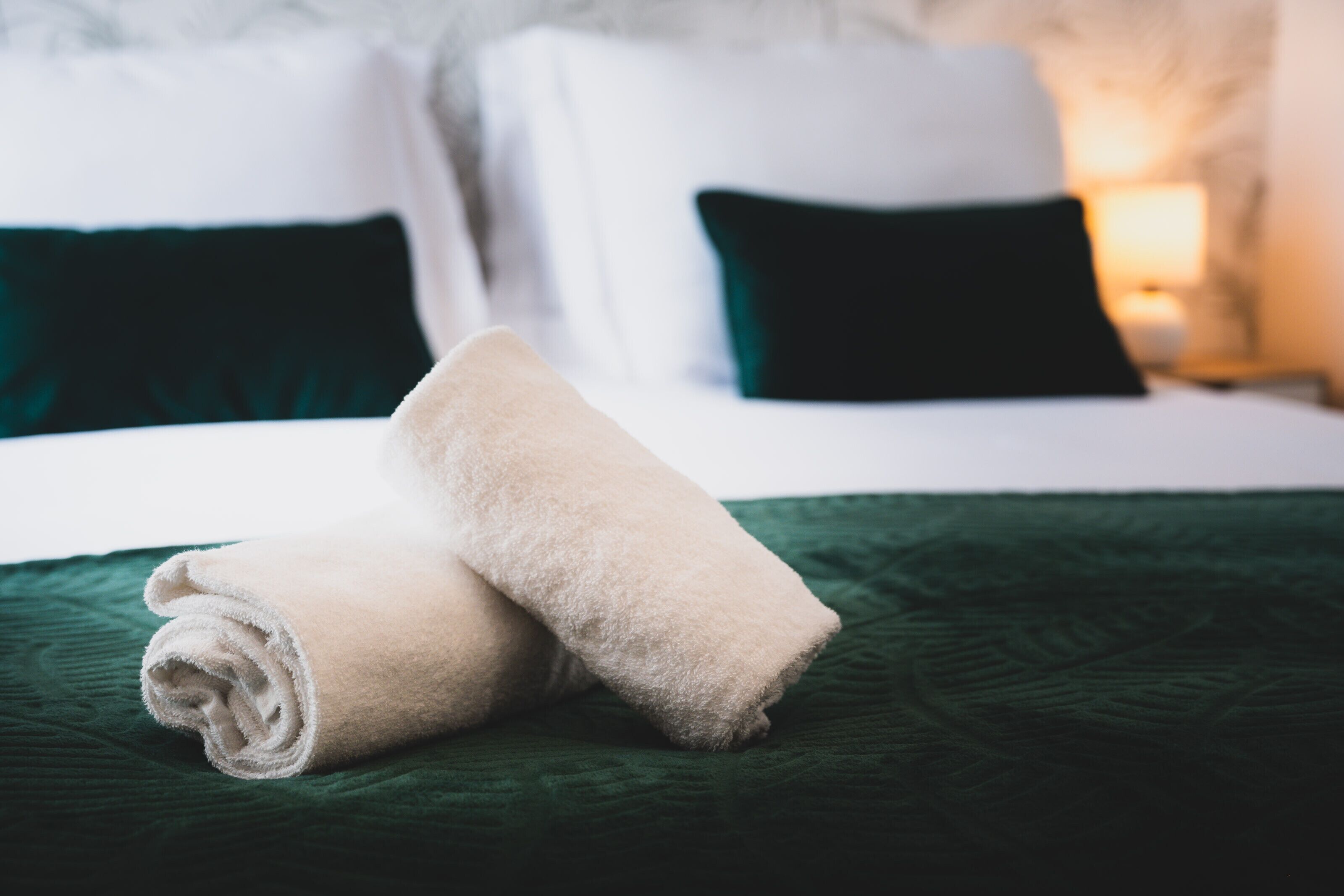 A neatly made bed with a green bedspread and two rolled towels.