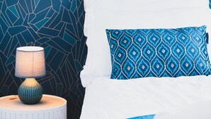 A neatly made bed with a blue blanket and a patterned pillow, a small round table with a lamp, and a geometric-patterned wall. - L'Art de Ville*** - 2  bedroom near the city center, terrace and garage (Annecy)