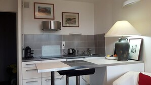 A compact kitchen with a white countertop, a coffee machine, a kettle, and a bowl of fruit. There are two framed pictures on the wall and a lamp on the right side. - Charming bright T2, balcony, parking, telework (Annecy)