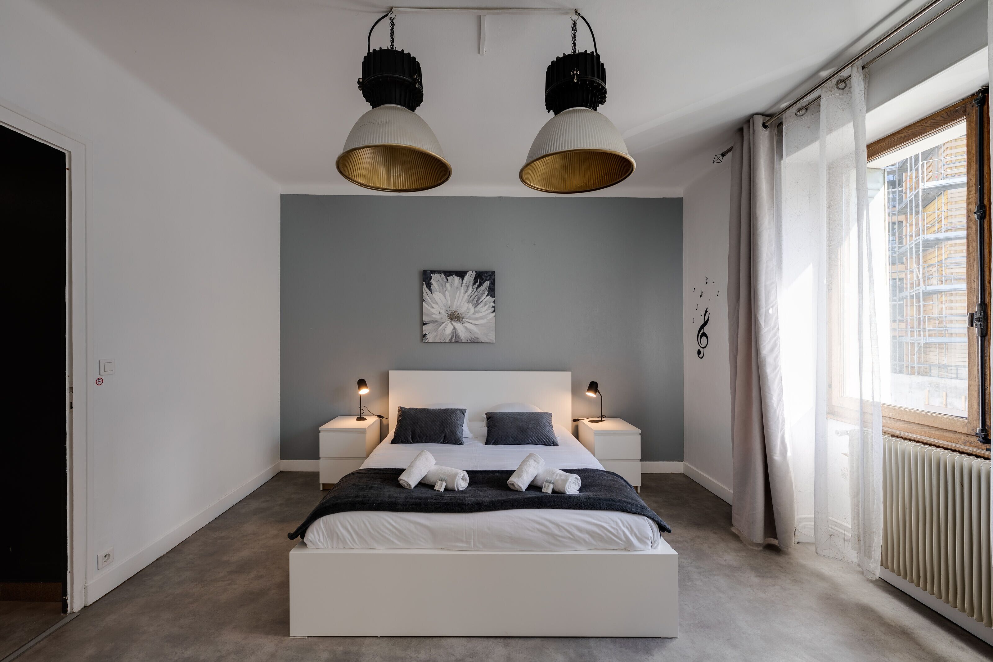 A modern bedroom with a large bed, two bedside tables with lamps, a ceiling light fixture, a radiator, and a window with curtains.