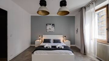 A modern bedroom with a large bed, two bedside tables with lamps, a ceiling light fixture, a radiator, and a window with curtains.
