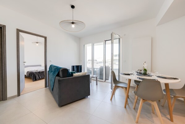 A modern living room with a sofa, dining table, and chairs. There is a balcony with a view.