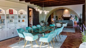 Un espace repas avec une table en verre entourée de chaises turquoise, un plafond en bois et une arche en pierre menant à la cuisine.