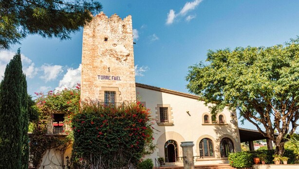 Un bâtiment historique doté d’une tour portant l’inscription « TORRE FAEL », avec une cour intérieure agrémentée d’une fontaine, entourée de verdure et surplombée d’un ciel dégagé.