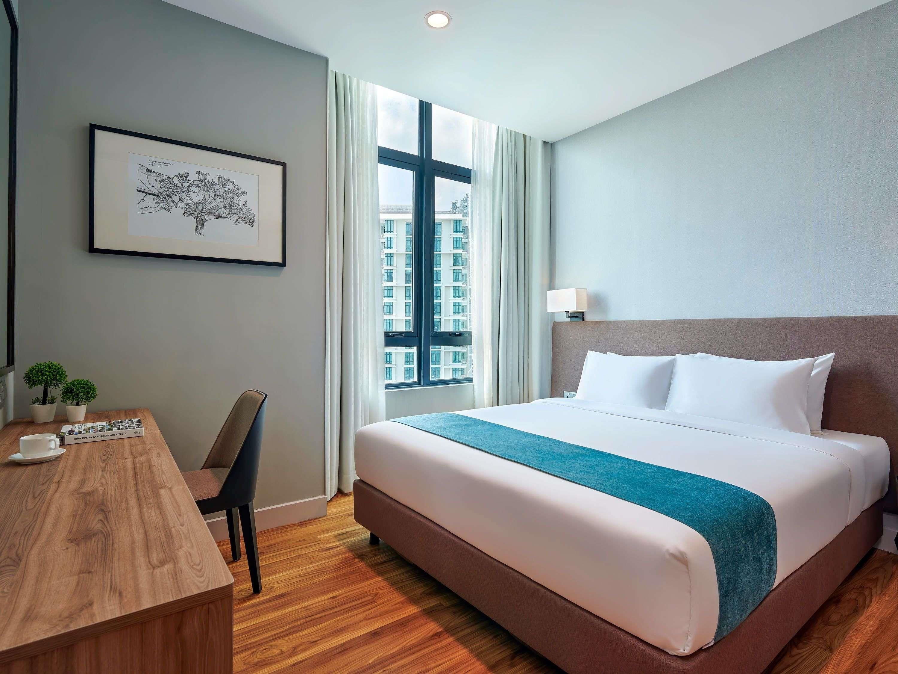 A hotel room with a large bed, a desk with a chair, a framed picture on the wall, and a window with a city view.