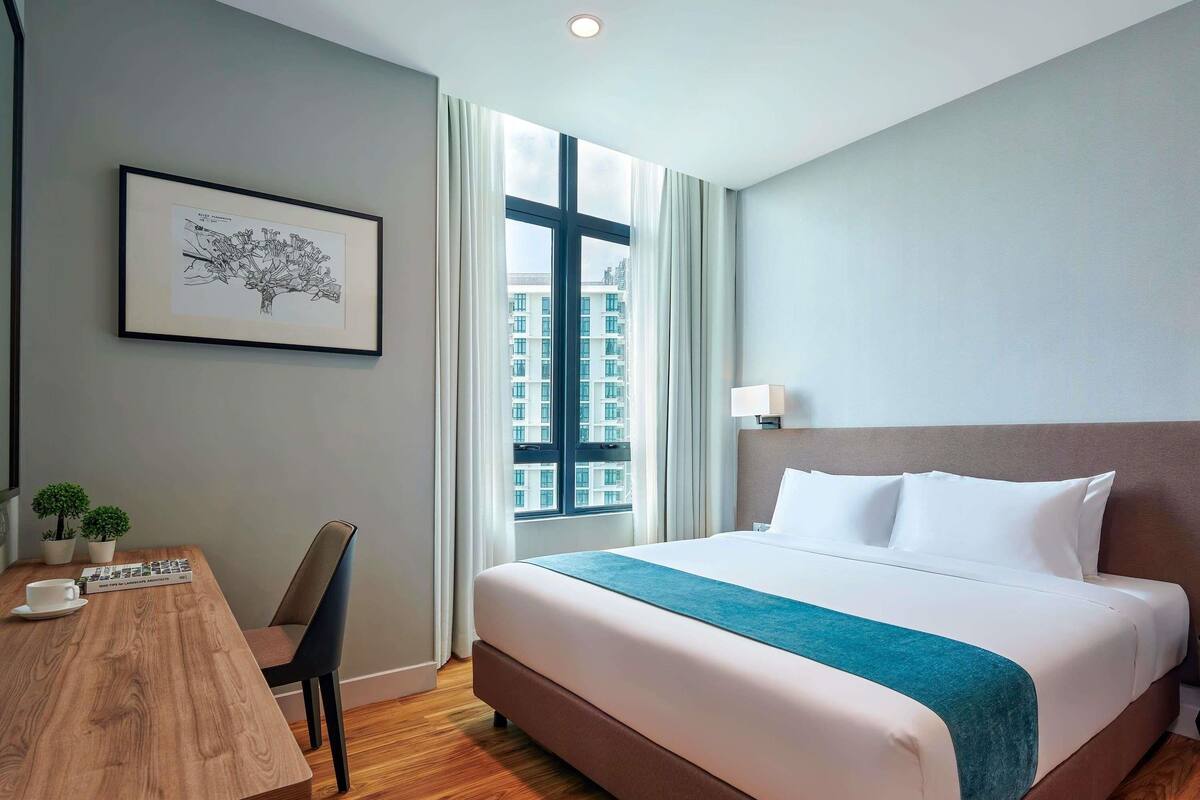Une chambre d’hôtel avec un grand lit, un bureau avec une chaise, un tableau encadré au mur et une fenêtre donnant sur la ville.