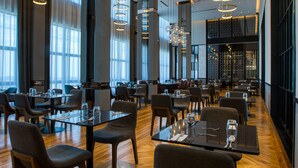 Een ruim restaurant met hoge plafonds, moderne hanglampen en een houten vloer. Tafels zijn netjes ingericht met stoelen, en grote ramen zorgen voor natuurlijk licht.