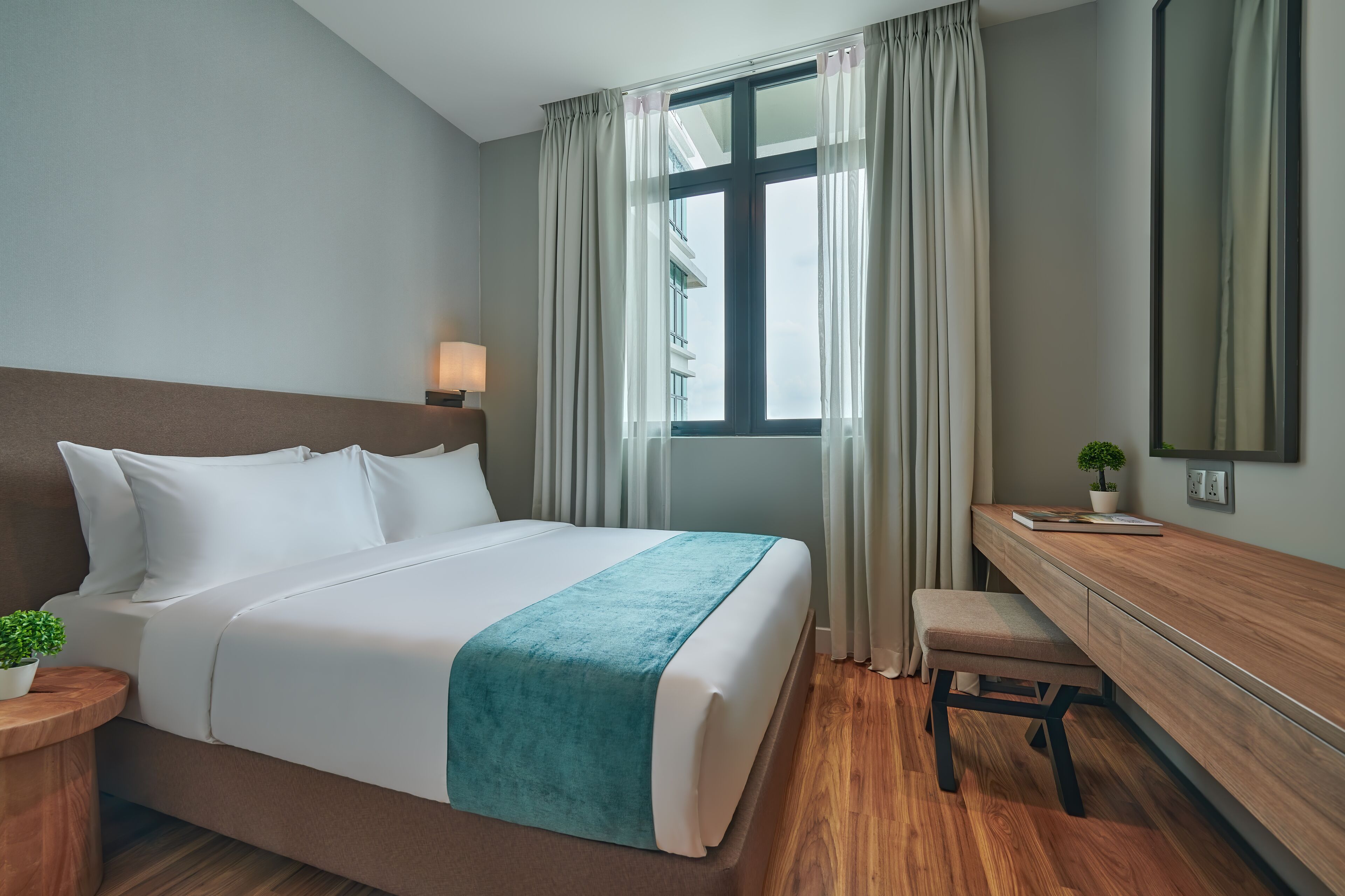 A hotel room with a bed, a desk, a chair, a potted plant, and a window with curtains.