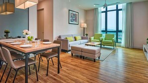 Une chambre d’hôtel moderne dotée d’un coin repas, d’un canapé, d’un fauteuil vert et d’une grande fenêtre donnant sur un paysage agréable.