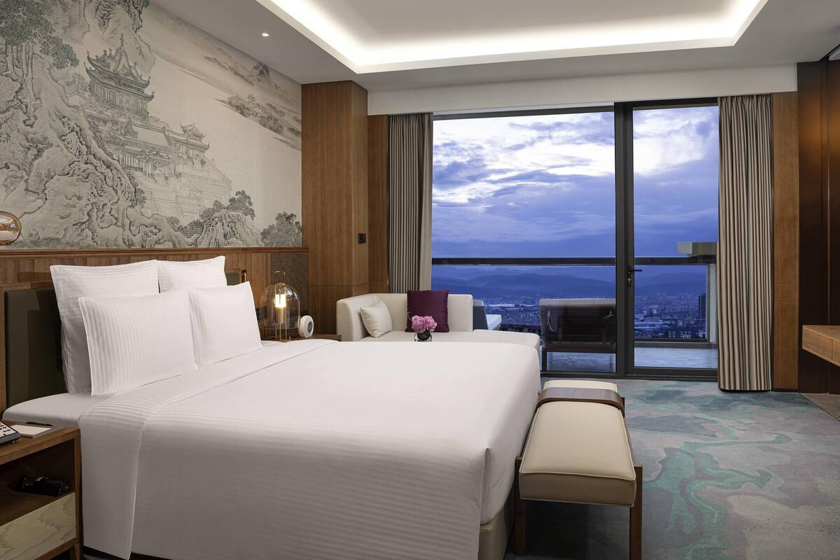 A modern hotel room with a large bed, a desk, and a view of the city through floor-to-ceiling windows.