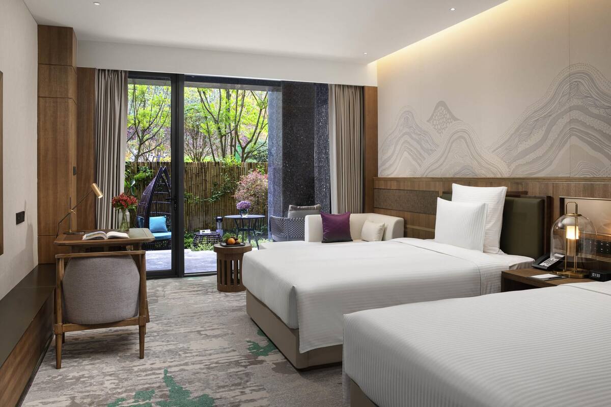 A hotel room with two beds, a desk, a chair, a small table with a vase, and a view of greenery outside.