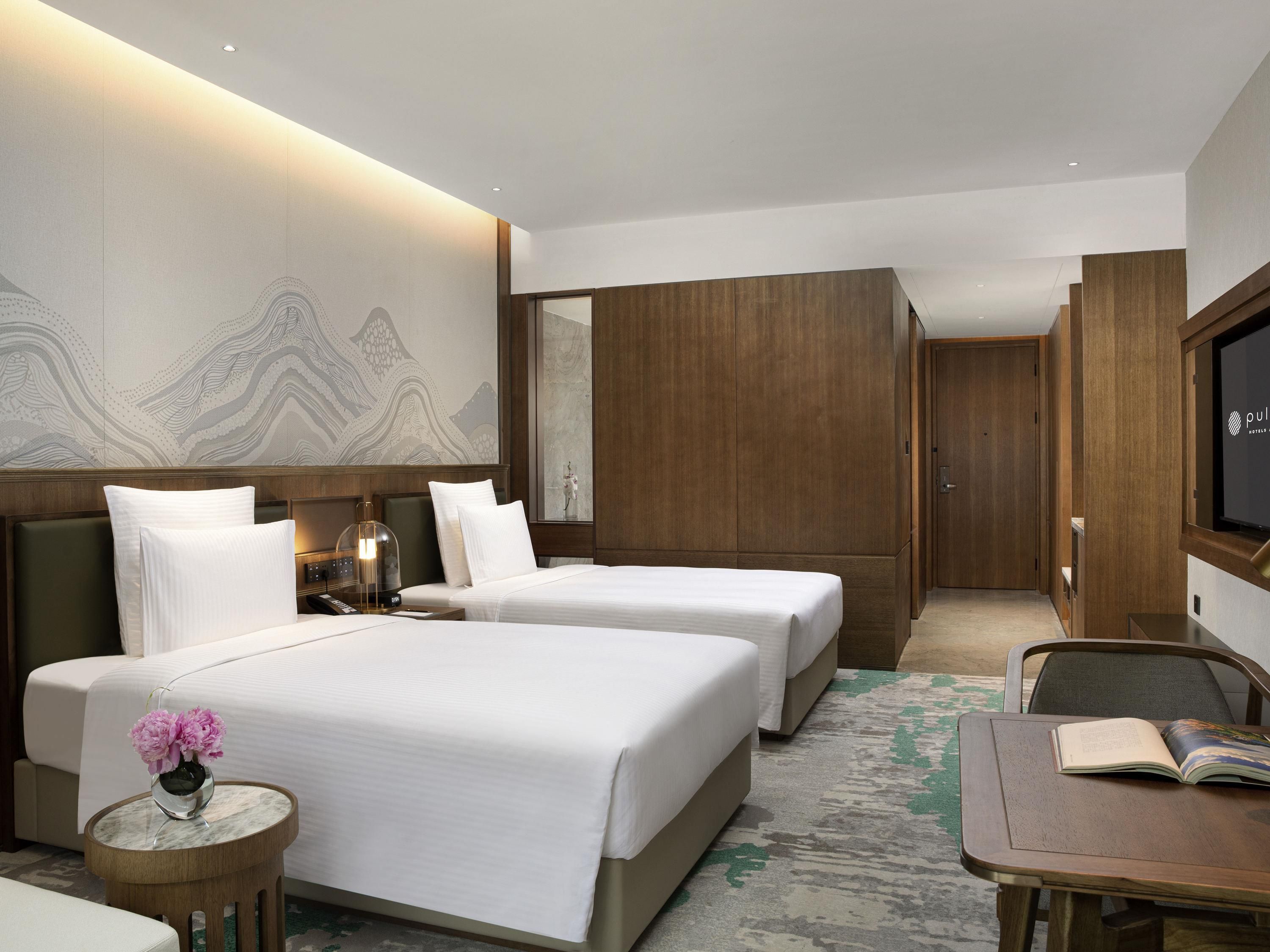 Una habitación de hotel con dos camas, una mesita con un jarrón de flores, un televisor y un espejo grande.