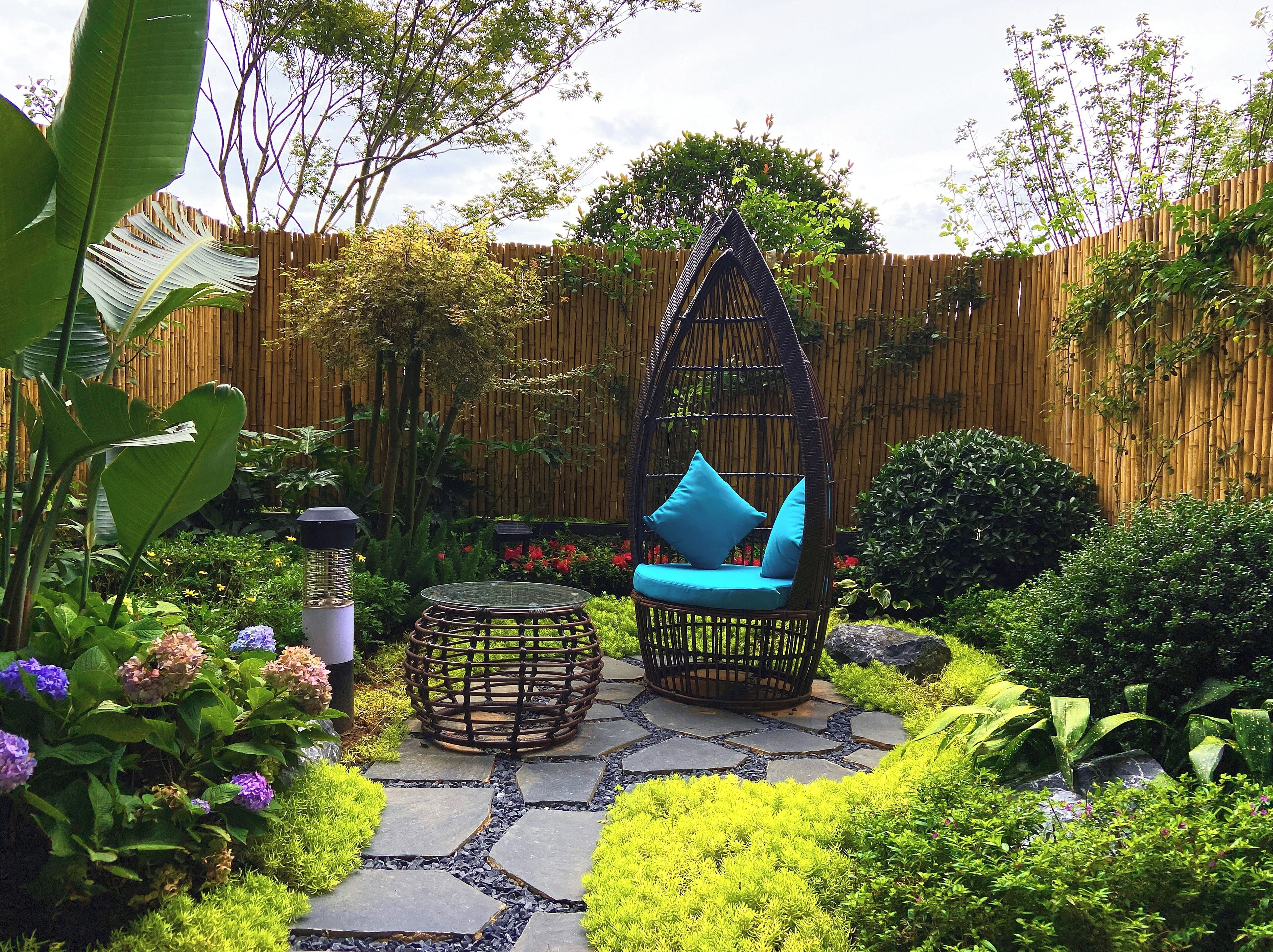 A garden with a wicker chair and a table, surrounded by greenery and a wooden fence.