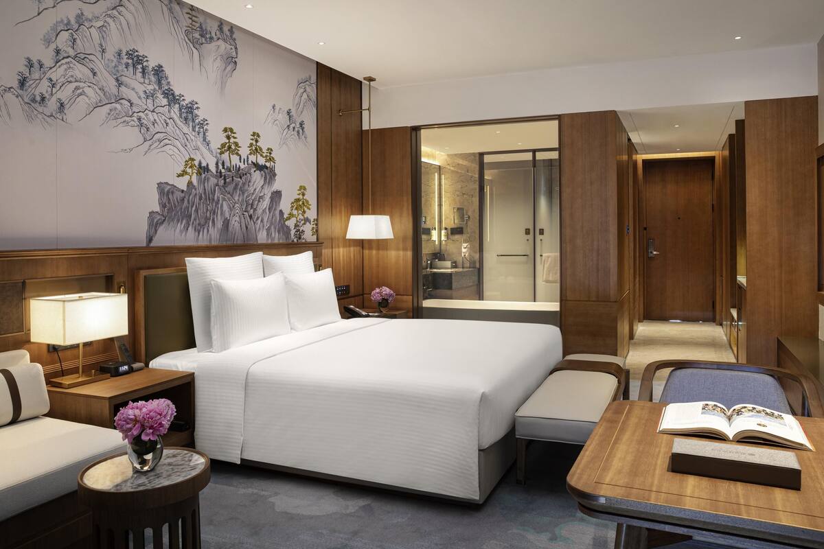 A modern hotel room with a large bed, bedside tables, a desk, and a bathroom visible through a glass door.