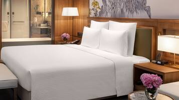 Une chambre d’hôtel avec un grand lit, une table de chevet avec une lampe, une petite table avec un téléphone et une salle de bain visible à travers une porte en verre.