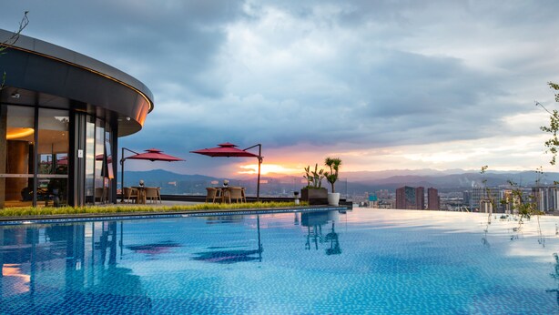A modern hotel with a pool, outdoor seating, and a view of the city skyline at sunset.