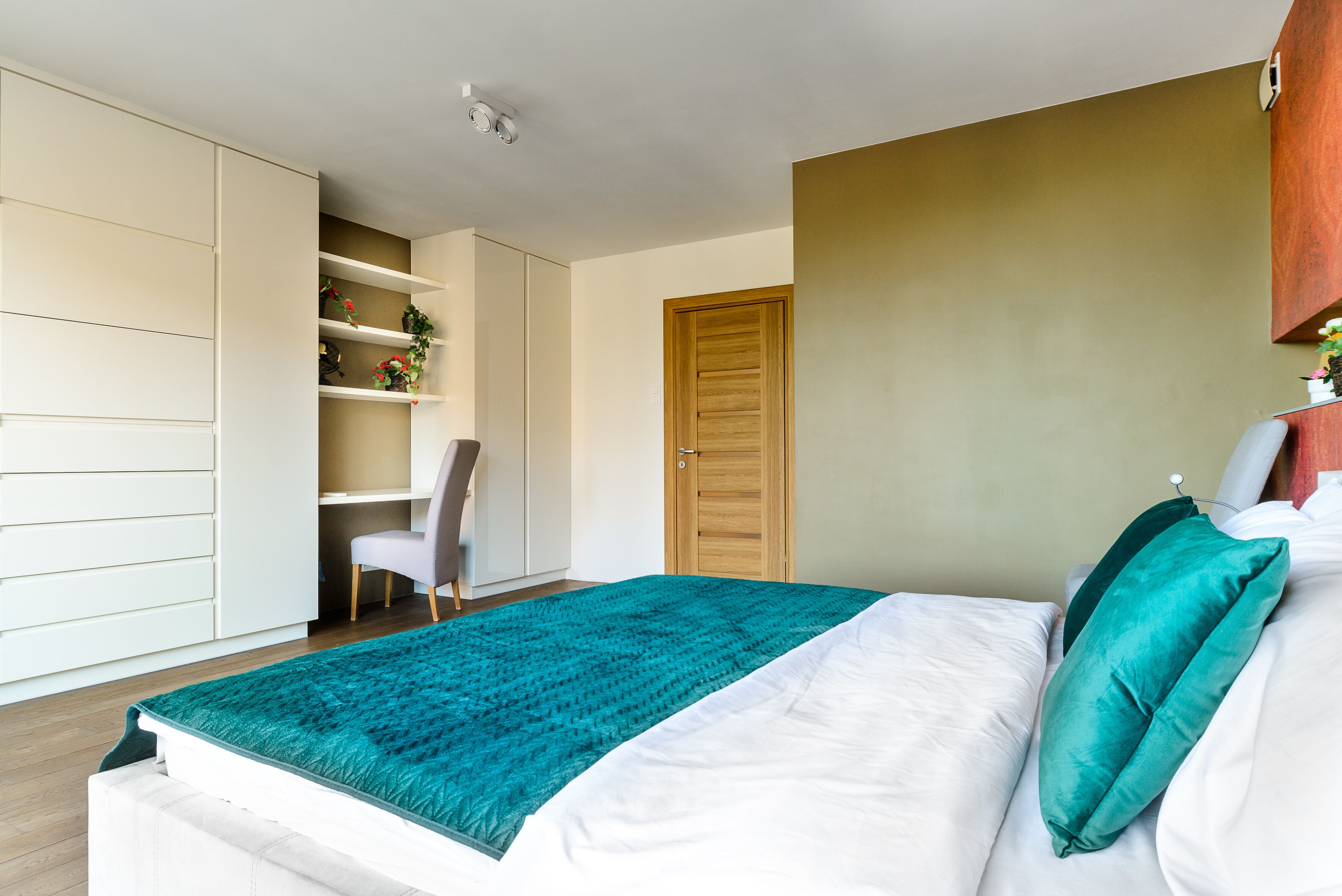 A bedroom with a bed, a teal blanket, a wooden door, and a white wardrobe.