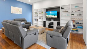 Una sala de estar moderna con sofás de cuero gris, una mesa de centro y un televisor de pantalla plana montado en una pared blanca. Se observan estantes con objetos decorativos y libros.