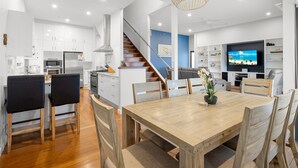 Una cocina moderna con armarios blancos, una mesa de comedor de madera y una escalera que lleva a una sala de estar con televisión.