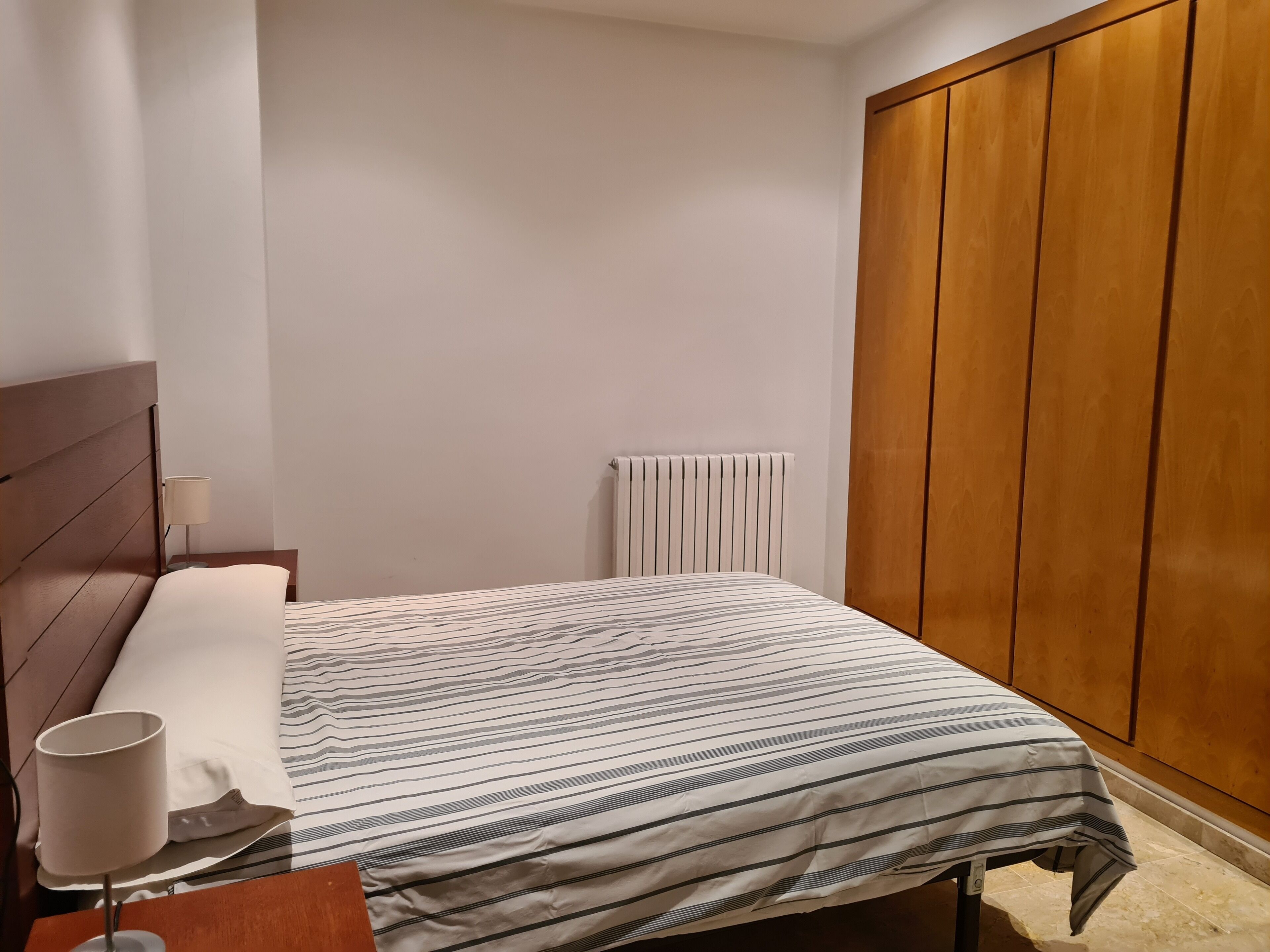 A bedroom with a bed, a nightstand, a lamp, a radiator, and a wooden wardrobe.