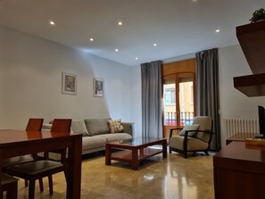 Una sala de estar con sofá, una mesa de centro, una silla y una mesa de comedor. Hay luces de techo y una ventana con cortinas.