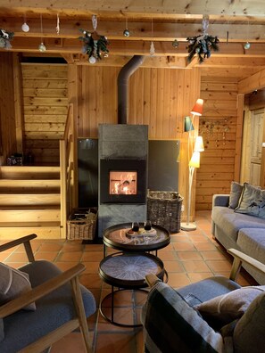 Ein gemütliches Blockhaus-Interieur mit Holzofen, einer Couch, einem runden Tisch mit Gläsern und einer Treppe.