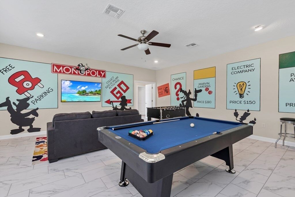 Une table de billard dans une pièce dotée d’un ventilateur de plafond, d’un canapé et de murs décorés d’affiches sur le thème du jeu de Monopoly et d’autres jeux de société.