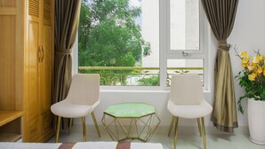 Een kamer met twee witte stoelen, een klein groen tafeltje, een raam met uitzicht op bomen en een potplant met gele bloemen.