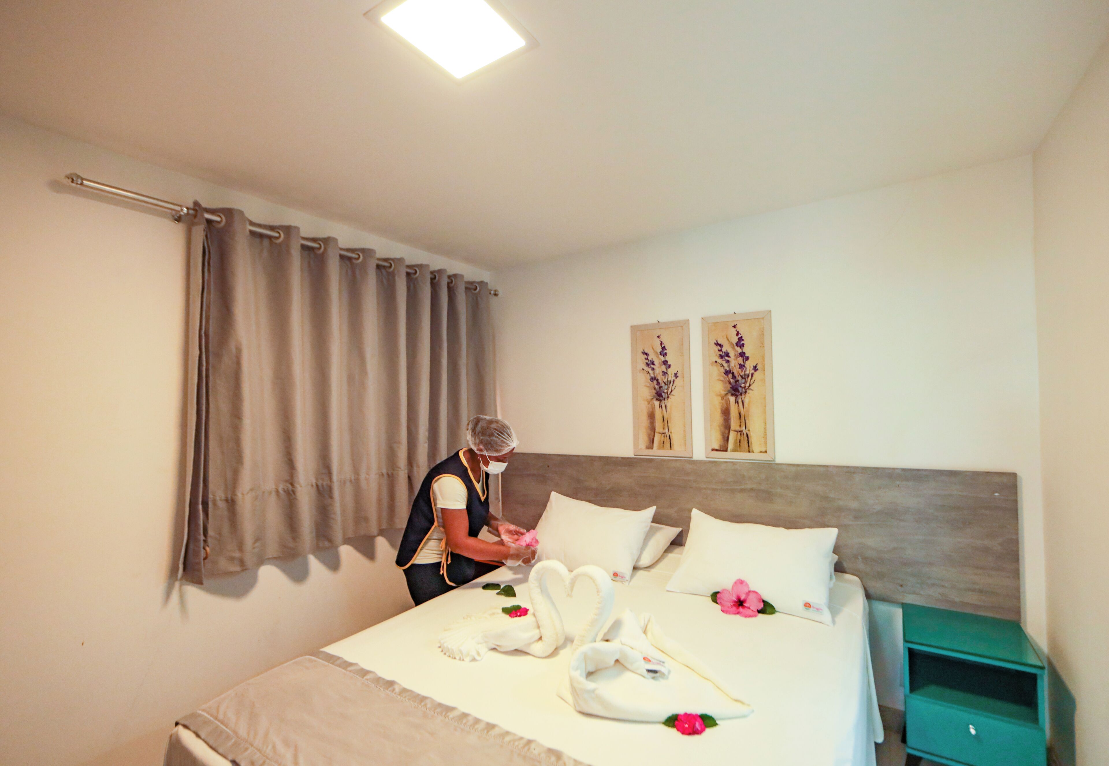 A hotel room with a neatly made bed, a green nightstand, and a person arranging pillows.
